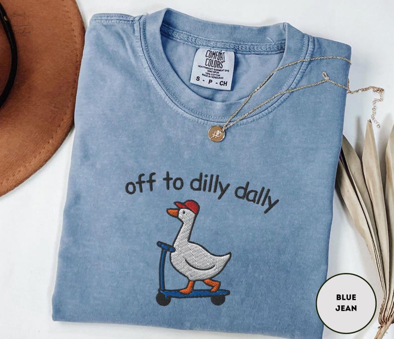 May include: A blue jean colored t-shirt with the phrase "off to dilly dally" embroidered above a cartoon goose riding a scooter. The goose wears a red hat. A gold necklace with a pendant is draped across the shirt.
