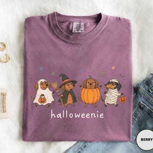 Op de afbeelding: Een bessenkleurig T-shirt met ronde hals en een afbeelding van vier teckels in Halloween-kostuums: een spook, een heks met een bezem, een pompoen en een mummie. Elke hond houdt een kleine oranje pompoen vast. De tekst "halloweenie" staat eronder gedrukt.