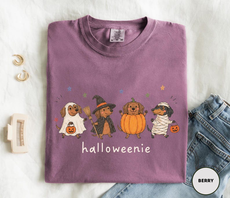 Op de afbeelding: Een bessenkleurig T-shirt met ronde hals en een afbeelding van vier teckels in Halloween-kostuums: een spook, een heks met een bezem, een pompoen en een mummie. Elke hond houdt een kleine oranje pompoen vast. De tekst "halloweenie" staat eronder gedrukt.