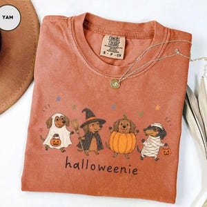 Op de afbeelding: Een yam-kleurig t-shirt met een afbeelding van vier teckels in Halloween-kostuums: een geest, een heks, een pompoen en een mummie. De tekst "halloweenie" staat onder de honden gedrukt. Het t-shirt heeft een Comfort Colors merklabel.
