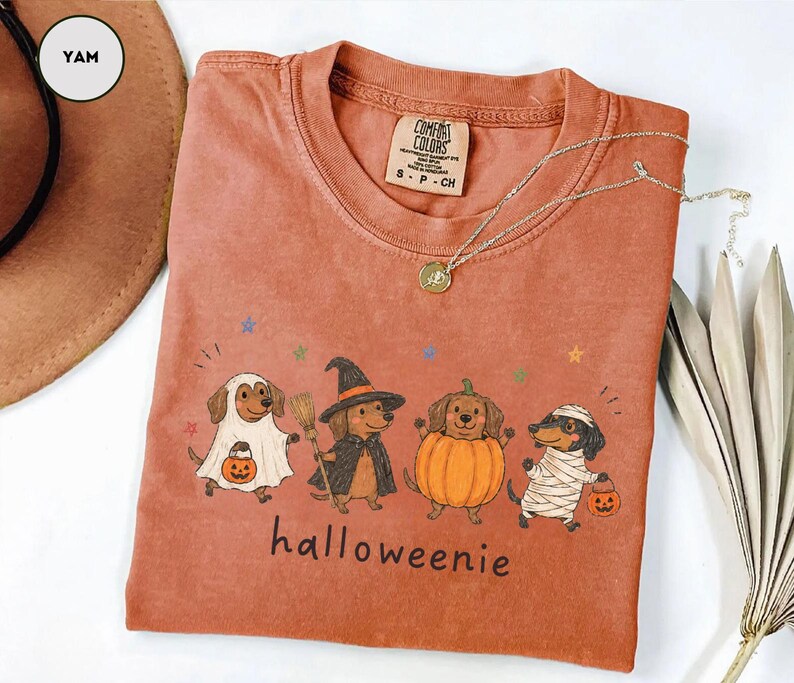 Op de afbeelding: Een yam-kleurig t-shirt met een afbeelding van vier teckels in Halloween-kostuums: een geest, een heks, een pompoen en een mummie. De tekst "halloweenie" staat onder de honden gedrukt. Het t-shirt heeft een Comfort Colors merklabel.