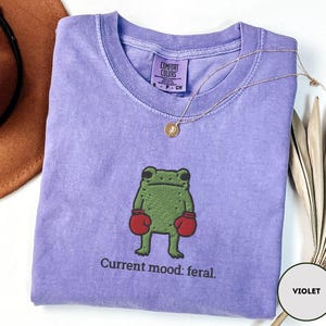May include: A violet Comfort Colors t-shirt featuring a green frog wearing red boxing gloves. The text "Current mood: feral" is below the frog. A gold necklace is draped across the shirt.