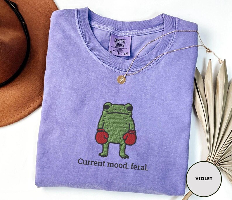 May include: A violet Comfort Colors t-shirt featuring a green frog wearing red boxing gloves. The text "Current mood: feral" is below the frog. A gold necklace is draped across the shirt.