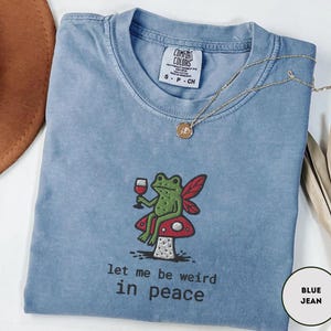 May include: A blue jean Comfort Colors t-shirt with a whimsical embroidered design. The design features a green frog with red wings, holding a glass of wine, sitting on a red and white mushroom. The text "let me be weird in peace" is below the design.
