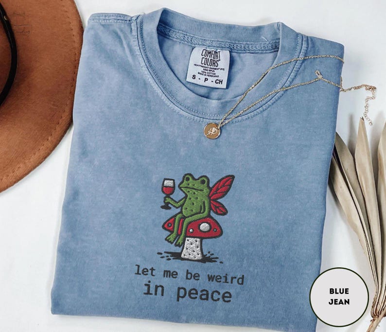 May include: A blue jean Comfort Colors t-shirt with a whimsical embroidered design. The design features a green frog with red wings, holding a glass of wine, sitting on a red and white mushroom. The text "let me be weird in peace" is below the design.