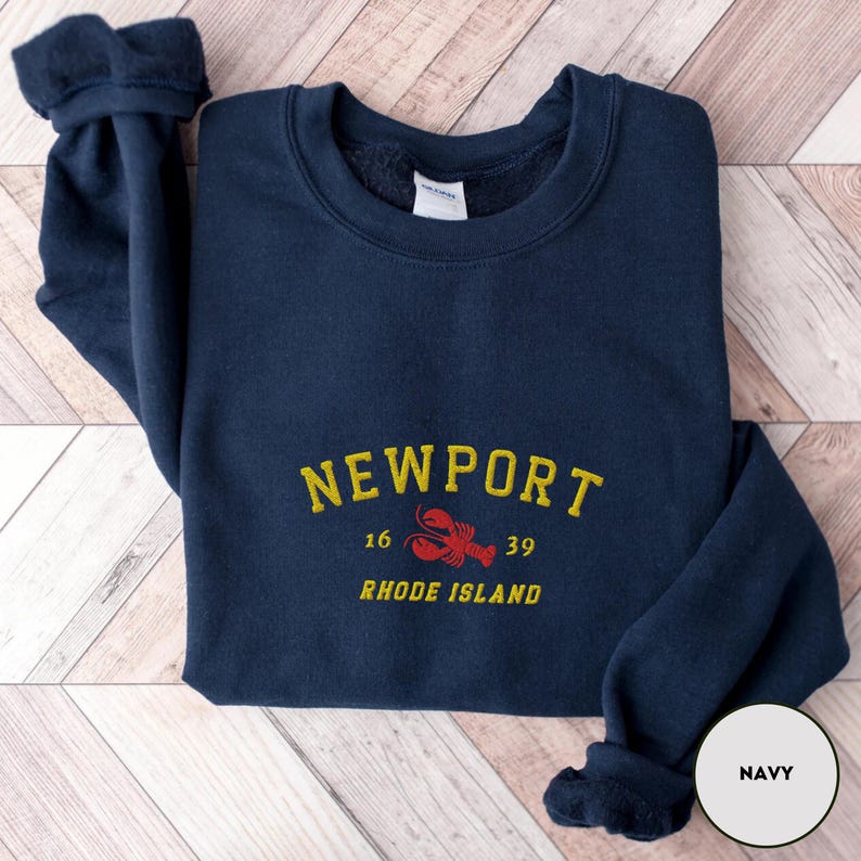 May include: Navy blue sweatshirt featuring the word "NEWPORT" in yellow arched text. Below the text is a red lobster graphic and the words "RHODE ISLAND". The sweatshirt has a crew neck and long sleeves.
