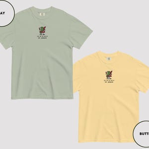 May include: Two short-sleeved t-shirts, one bay green and one butter yellow, each featuring an embroidered design of a frog holding a glass of wine. The text "let me be weird in peace" is below the frog. Casual wear.