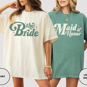 May include: Two oversized t-shirts, one ivory and one light green. The ivory shirt has the text "the Bride" in a retro font. The light green shirt says "Maid of Honor". Both shirts are short-sleeved.