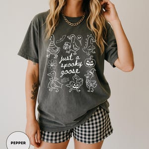 May include: Dark grey t-shirt with a white graphic of cartoon geese in Halloween costumes and the text "just a spooky goose". The shirt is paired with black and white checkered shorts. A gold chain necklace is visible.