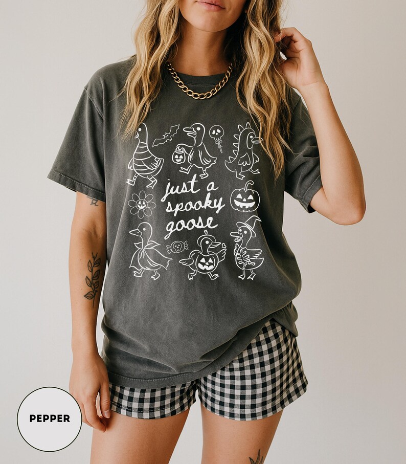 May include: Dark grey t-shirt with a white graphic of cartoon geese in Halloween costumes and the text "just a spooky goose". The shirt is paired with black and white checkered shorts. A gold chain necklace is visible.