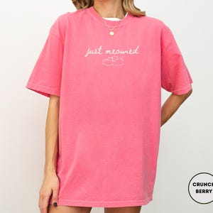 May include: A short-sleeved, oversized, pink t-shirt with the phrase "just meowied" in cursive script and a small cat illustration. The shirt is a solid color and has a relaxed fit. The brand name "Crunch Berry" is in a circle on the bottom right.
