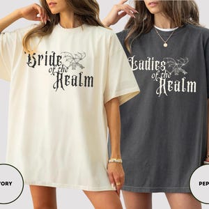 May include: Two oversized t-shirts, one ivory and one pepper, featuring the text "Bride of the Realm" and "Ladies of the Realm" in a gothic font with a dragon illustration. The shirts have short sleeves and a relaxed fit.
