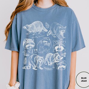 Op de afbeelding: Blauw spijkerbroekkleurig T-shirt met een witte illustratie van wasberen in verschillende poses. Het ontwerp bevat verschillende wasberen, sommige met bladaccenten.