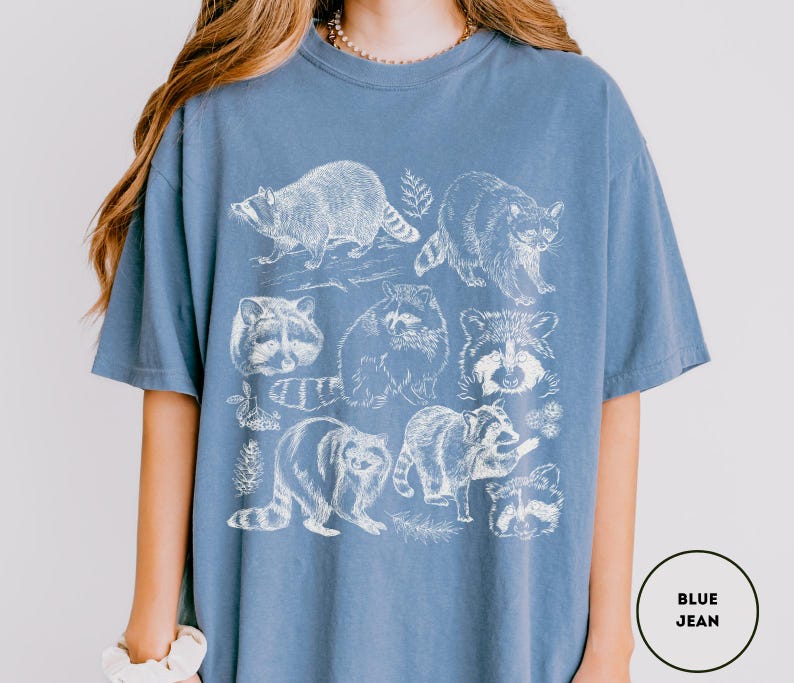 Op de afbeelding: Blauw spijkerbroekkleurig T-shirt met een witte illustratie van wasberen in verschillende poses. Het ontwerp bevat verschillende wasberen, sommige met bladaccenten.