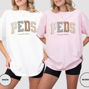 May include: Two oversized t-shirts, one white and one pink, featuring the text "PEDS nurse" in a collegiate-style design. The letters have a floral pattern. The shirts are worn with black shorts.