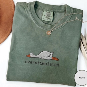 May include: A moss green t-shirt with a crew neck and short sleeves. The shirt features an embroidered graphic of a duck with the word "overstimulated" below it. A gold necklace with a pendant is draped on the shirt.