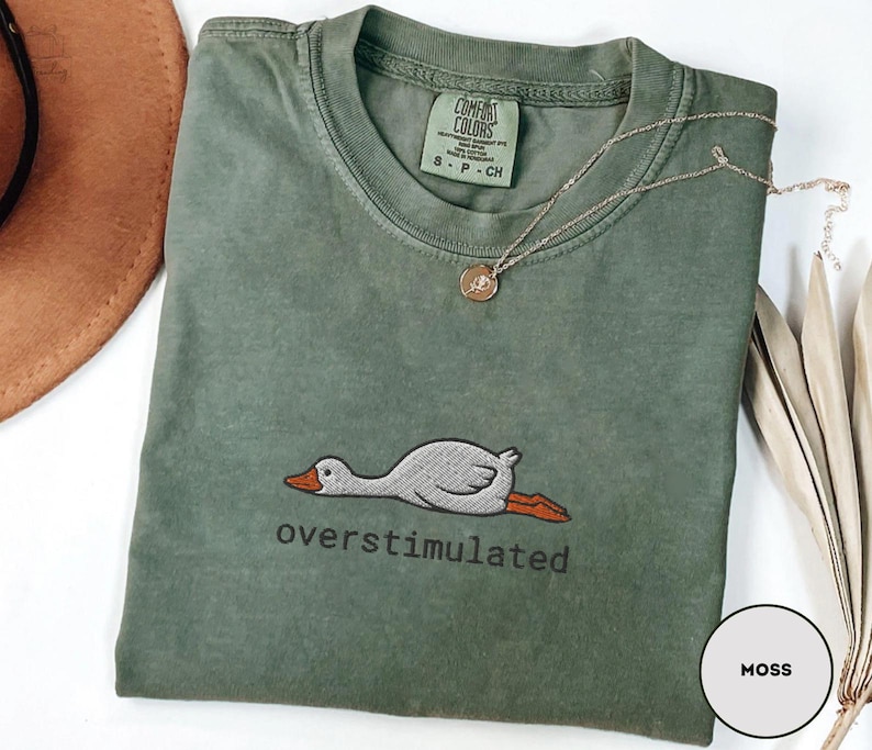 May include: A moss green t-shirt with a crew neck and short sleeves. The shirt features an embroidered graphic of a duck with the word "overstimulated" below it. A gold necklace with a pendant is draped on the shirt.