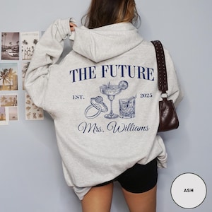 Pode incluir: Um moletom cinza com o texto "The Future Est. 2025 Mrs. Williams" estampado na frente. O design inclui um copo de coquetel, um anel e um cora&ccedil;&atilde;o.