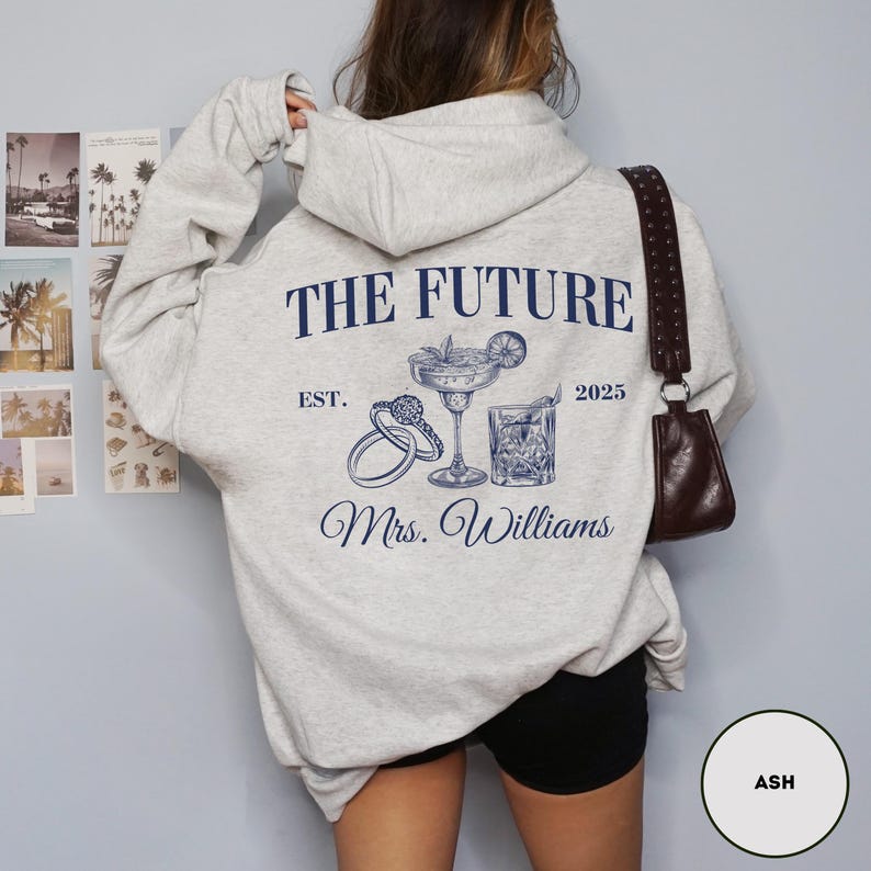 Pode incluir: Um moletom cinza com o texto "The Future Est. 2025 Mrs. Williams" estampado na frente. O design inclui um copo de coquetel, um anel e um cora&ccedil;&atilde;o.