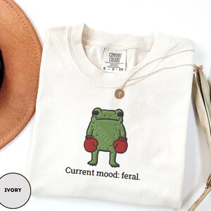 May include: Ivory-colored t-shirt with an embroidered green frog wearing red boxing gloves. The text "Current mood: feral" is below the frog. A gold necklace and a brown hat are also visible.