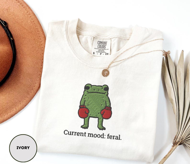 May include: Ivory-colored t-shirt with an embroidered green frog wearing red boxing gloves. The text "Current mood: feral" is below the frog. A gold necklace and a brown hat are also visible.