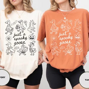 May include: Ivory and yam-colored t-shirts with a Halloween-themed graphic. The graphic features cartoon geese in costumes, a pumpkin, and the text "just a spooky goose". The shirts are oversized and casual.