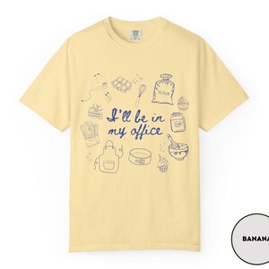 May include: Banana-colored t-shirt with a blue illustration of baking tools and the phrase "I'll be in my office." The design includes a mixer, eggs, a whisk, a bag of flour, cupcakes, and an apron.