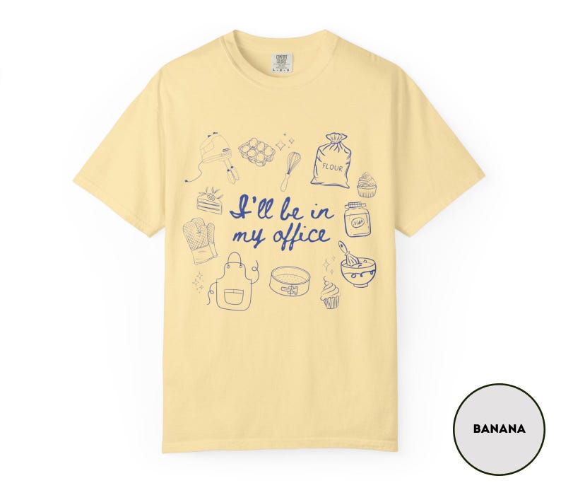 May include: Banana-colored t-shirt with a blue illustration of baking tools and the phrase "I'll be in my office." The design includes a mixer, eggs, a whisk, a bag of flour, cupcakes, and an apron.