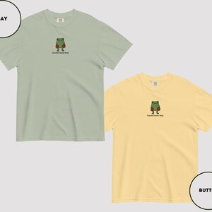 May include: Two short-sleeved t-shirts, one bay green and one butter yellow, each featuring an embroidered frog wearing boxing gloves. The text "Current mood: fend." is below the frog. The shirts are displayed on a light gray background.