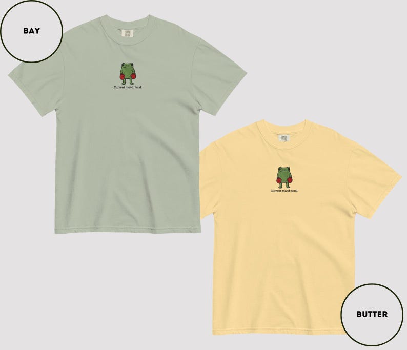 May include: Two short-sleeved t-shirts, one bay green and one butter yellow, each featuring an embroidered frog wearing boxing gloves. The text "Current mood: fend." is below the frog. The shirts are displayed on a light gray background.