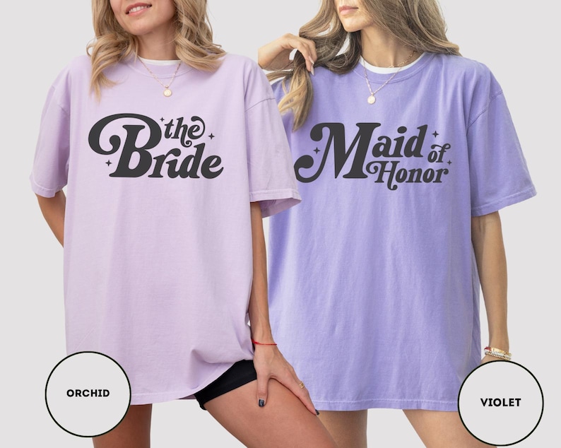 May include: Two oversized t-shirts are shown, one orchid and one violet. The orchid shirt features the text "the Bride" in a black, retro font. The violet shirt says "Maid of Honor" in a similar style. Both are short-sleeved.