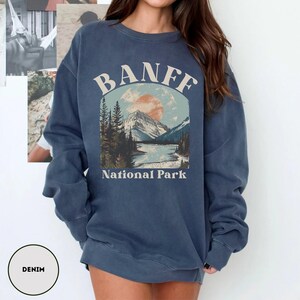 May include: A blue denim sweatshirt with a vintage-style graphic of a mountain landscape and the text "Banff National Park".