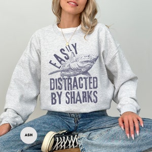 May include: A gray sweatshirt with the text "Easily Distracted By Sharks" and a graphic of a shark. The sweatshirt is being modeled by a person wearing blue jeans.