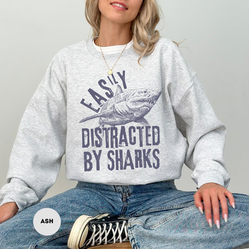 May include: A gray sweatshirt with the text "Easily Distracted By Sharks" and a graphic of a shark. The sweatshirt is being modeled by a person wearing blue jeans.
