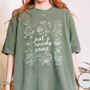 May include: Moss green t-shirt with a white graphic of cartoon geese in Halloween costumes and the text "just a spooky goose". The shirt is made of a soft material.
