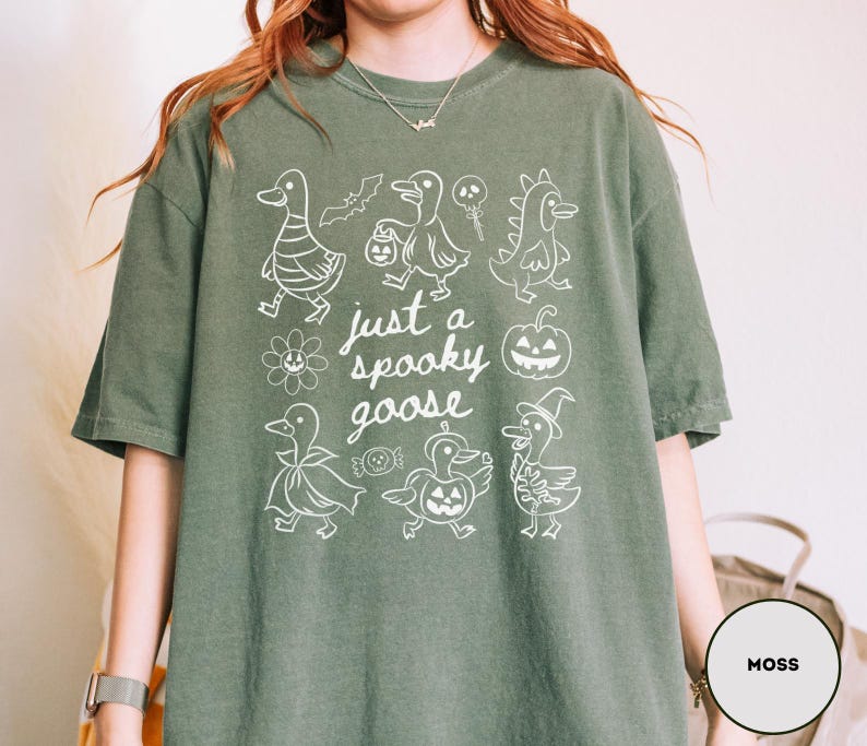 May include: Moss green t-shirt with a white graphic of cartoon geese in Halloween costumes and the text "just a spooky goose". The shirt is made of a soft material.