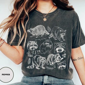 Op de afbeelding: Donkergrijs t-shirt met een witte illustratie van wasberen in verschillende poses, met wat bladeren. Het t-shirt heeft een vintage, versleten uitstraling. Het woord "Pepper" staat in een cirkel.