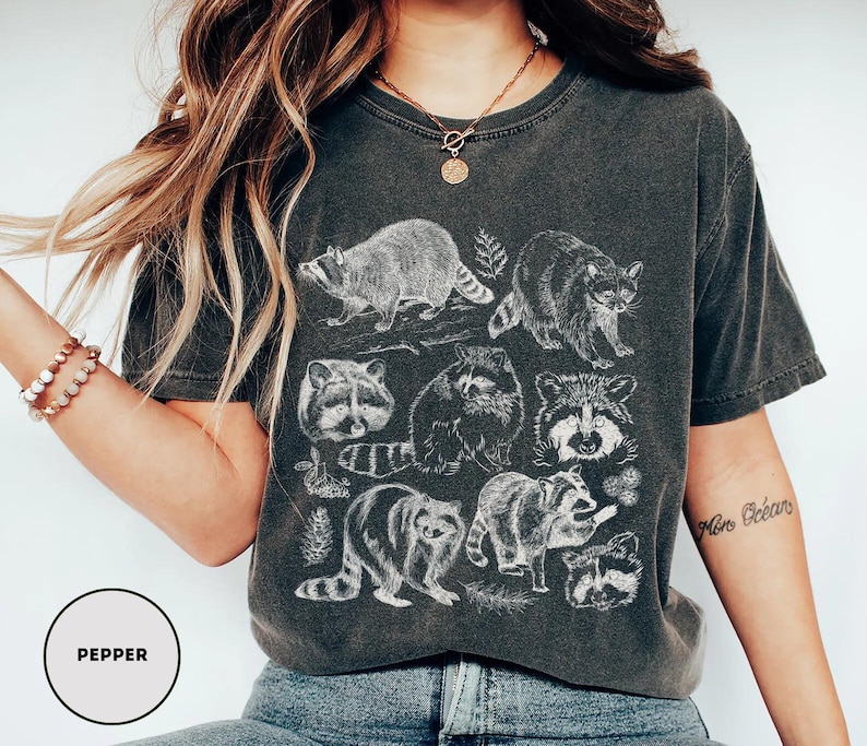 Op de afbeelding: Donkergrijs t-shirt met een witte illustratie van wasberen in verschillende poses, met wat bladeren. Het t-shirt heeft een vintage, versleten uitstraling. Het woord "Pepper" staat in een cirkel.