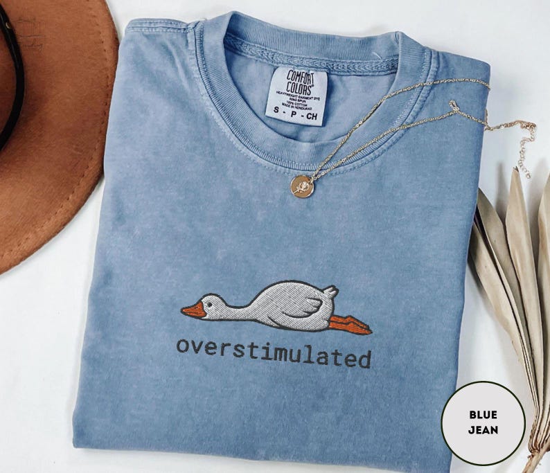 May include: A blue jean-coloured t-shirt with the word "overstimulated" embroidered below a cartoon goose. The shirt has a crew neck and short sleeves. A gold necklace with a pendant is draped across the shirt.