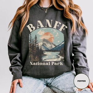 May include: A dark gray sweatshirt with a vintage-style graphic of a mountain landscape and the text "Banff National Park".