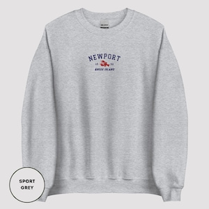 May include: A light grey sweatshirt featuring the word "NEWPORT" in navy blue, with a red lobster graphic. Below the lobster is the text "1639 RHODE ISLAND". The sweatshirt is labelled "SPORT GREY".