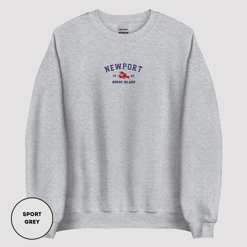 May include: A light grey sweatshirt featuring the word "NEWPORT" in navy blue, with a red lobster graphic. Below the lobster is the text "1639 RHODE ISLAND". The sweatshirt is labelled "SPORT GREY".