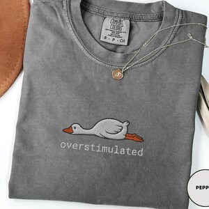May include: A grey Comfort Colors t-shirt featuring the word "overstimulated" embroidered beneath a cartoon goose. A silver necklace with a pendant lies on the shirt. The t-shirt is size small.