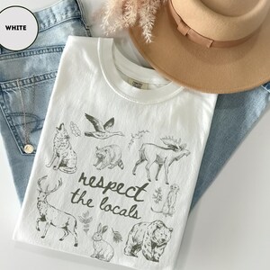 May include: White t-shirt with a crew neck featuring a graphic of various animals and the text "respect the locals". The shirt is laid on top of blue jeans and next to a tan hat.
