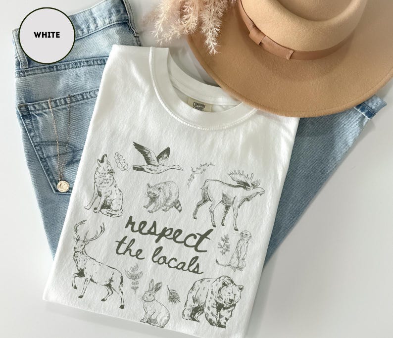 May include: White t-shirt with a crew neck featuring a graphic of various animals and the text "respect the locals". The shirt is laid on top of blue jeans and next to a tan hat.