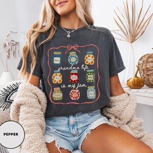 Peut inclure: T-shirt gris fonc&eacute; avec un motif de pots de confiture et le texte "grandma life is my jam". Le motif pr&eacute;sente des pots avec divers motifs de fruits et de fleurs, encadr&eacute;s par une bordure rose.