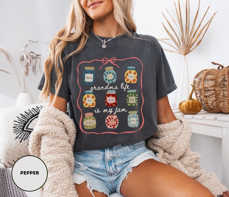 Peut inclure: T-shirt gris fonc&eacute; avec un motif de pots de confiture et le texte "grandma life is my jam". Le motif pr&eacute;sente des pots avec divers motifs de fruits et de fleurs, encadr&eacute;s par une bordure rose.