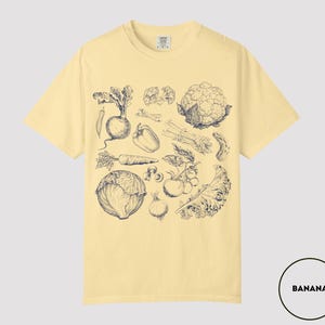 May include: A light yellow t-shirt featuring a detailed black and white illustration of various vegetables, including a carrot, cauliflower, and tomatoes. The shirt has a crew neck and short sleeves. The word "BANANA" is in a white circle in the bottom right corner.