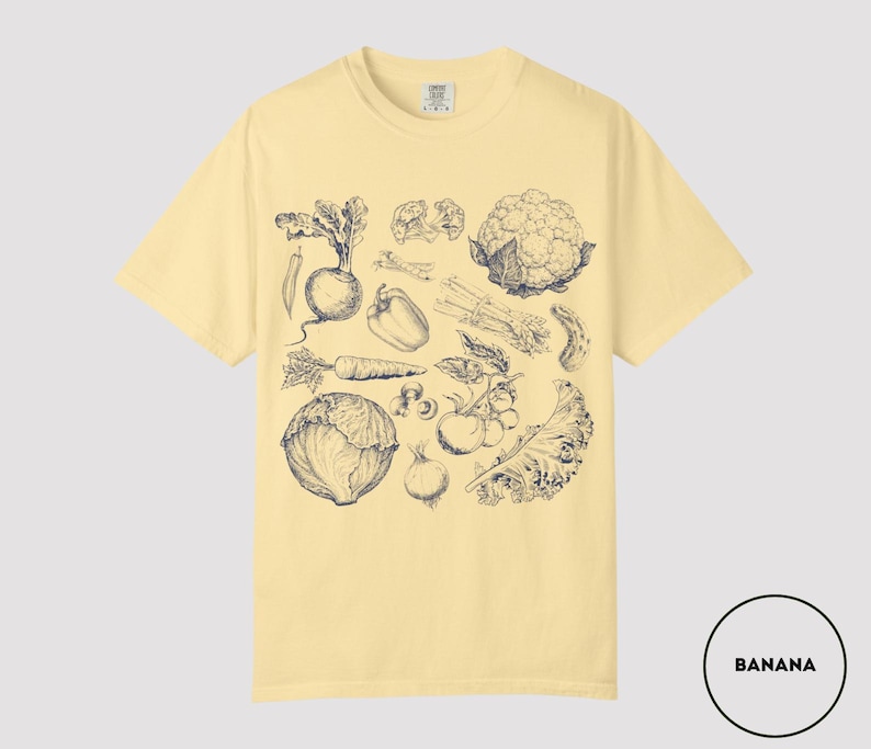 May include: A light yellow t-shirt featuring a detailed black and white illustration of various vegetables, including a carrot, cauliflower, and tomatoes. The shirt has a crew neck and short sleeves. The word "BANANA" is in a white circle in the bottom right corner.