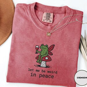 May include: Crimson-colored t-shirt with a crew neck. The shirt features an embroidered design of a green frog with fairy wings, holding a wine glass, sitting on a mushroom. The text "let me be weird in peace" is below the frog.